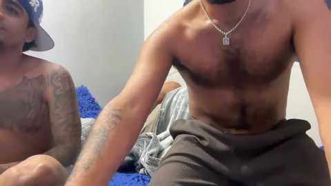 Video of two shirtless men in a bedroom; one with tattoos, the other with a necklace, sitting on a blue blanket.