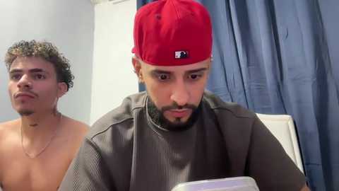 Video of a shirtless man with curly hair and a man in a red cap and grey shirt, both in a medical setting.