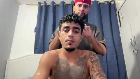 Video of two men in a bathroom: one shirtless with dark curly hair and a tattoo, the other in a red cap, adjusting his hair.