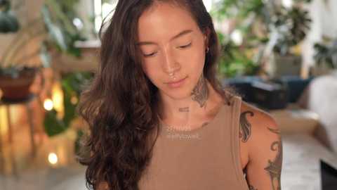 Video of a young woman with long, wavy brown hair, wearing a sleeveless tank top, standing in a bright, plant-filled room with wooden furniture. She has tattoos on her neck and shoulders.