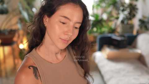 Video of a young woman with long, wavy brown hair, wearing a beige sleeveless top, with a nose ring, in a warmly lit, indoor setting featuring green plants and a white couch.
