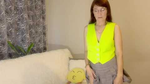 Video of a middle-aged woman with glasses, straight brown hair, and fair skin, wearing a bright yellow vest over a grey skirt, sitting on a white couch with a stuffed animal, in a beige room with floral curtains.