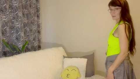 A young woman with long brown hair and glasses, wearing a neon yellow top and plaid skirt, stands in a cozy living room with a white sofa and floral curtains.