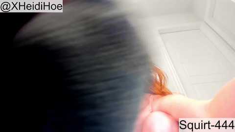 A blurred video shows a close-up of a person's hand with long, light brown hair, possibly a woman, in a bathroom setting. The background features white walls and a door. Text in the corners reads \"@XHeidiHo\" and \"Squirt-644.\