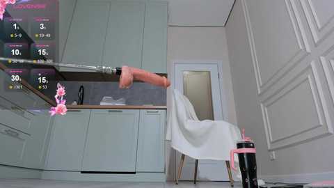 Video of a white kitchen with pastel cabinets, a hanging dildo, a pink chair, and a pink umbrella.
