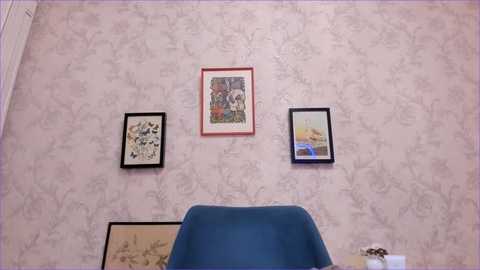 A video of a small, cozy room with a pastel, floral-patterned wallpaper. Three framed art pieces hang on the wall: a colorful landscape, a black-and-white sketch, and a botanical print. A blue chair is positioned centrally in the foreground.