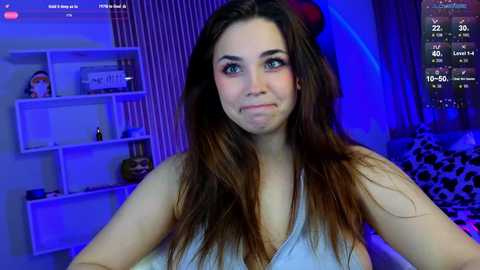 Video of a young woman with long, straight brown hair, wearing a sleeveless top, smiling in a dimly-lit room with a white bookshelf and a leopard-print blanket in the background.