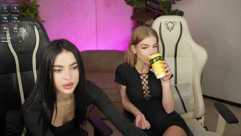 Video of two women in a modern room. One, with long black hair, wears a black dress, drinking a drink. The other, with light brown hair, sits in a gaming chair, holding a yellow cup, looking bored.