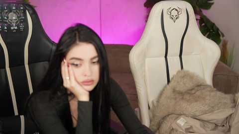 Video of a woman with long black hair and fair skin, wearing a black top, resting her head on her hand, in a modern living room with white gaming chairs and a cozy blanket.