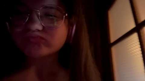 A dimly lit video of a young Asian woman with long hair, wearing clear glasses and a pink wireless headset, with a warm-toned, textured wall in the background.
