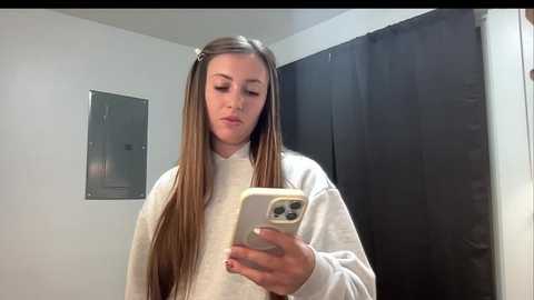 A video of a young woman with long, straight brown hair, wearing a white turtleneck sweater, taking a selfie in a dimly lit bathroom with a black shower curtain.
