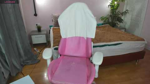 Video of a pink recliner chair with a white umbrella, set in a pastel-pink room with a wooden floor, featuring a bed with green and white bedding, a small gray nightstand, and a potted plant.