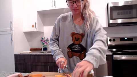 Video of a blonde woman in glasses, wearing a gray sweatshirt with a teddy bear graphic, cutting fruit on a kitchen island. Background features modern stainless steel appliances, white cabinets, and a vase with flowers.