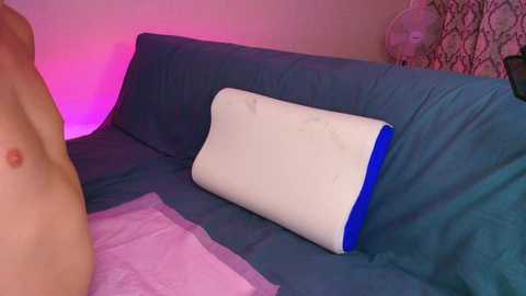 A video showing a nude person lying on a blue bedspread, with a white and blue pillow on their chest. Pink and purple lighting casts a soft glow on the scene.