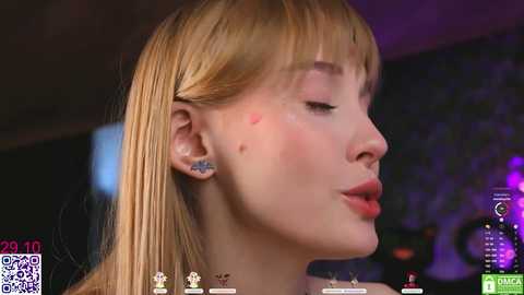 Video of a fair-skinned, blonde woman with bangs, wearing minimal makeup, a small butterfly tattoo on her cheek, and a necklace. Background is blurred with purple and blue hues.