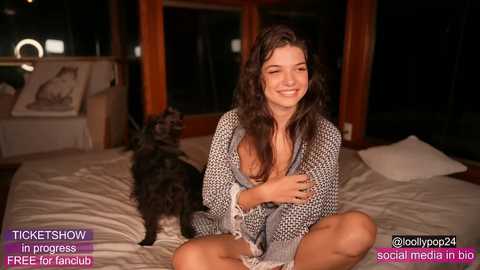 Video of a young woman with long dark hair, smiling, wearing a patterned cardigan, sitting on a bed with a black dog.
