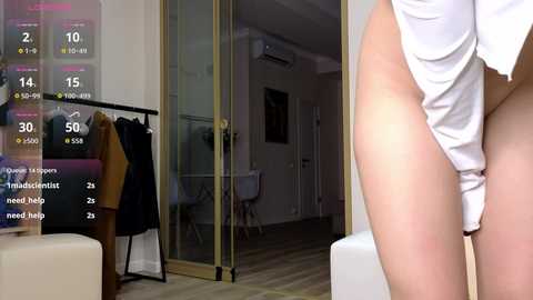 Video of a woman in white lingerie, standing in a modern apartment with wooden floors and minimalist decor. A digital display with weather and transport info is visible on the left.