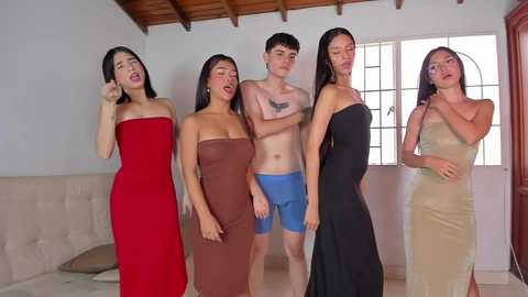 Video of five young women, four Asian and one Caucasian, dressed in tight, strapless dresses in red, brown, blue, black, and beige, standing in a modern living room with wooden ceiling beams and large windows.
