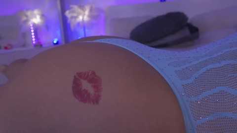 Media: Video of a person's lower back and buttocks, featuring a red lipstick kiss tattoo. They wear light blue mesh lingerie, lying on a bed with purple lighting and palm tree lampshades in the background.