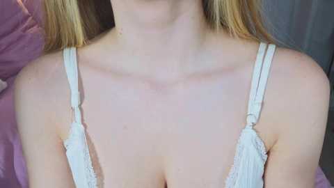 Video of a fair-skinned woman with long blonde hair, wearing a white lace bra, showing cleavage. She is seated on a purple pillow against a dark background.