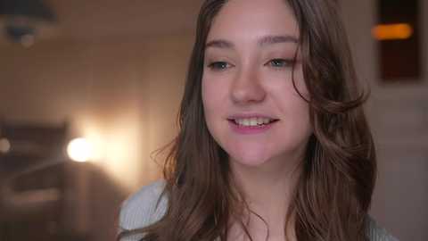 Video of a smiling young woman with long brown hair, wearing a light grey sweater, in a softly lit, warmly colored room with blurred background elements.