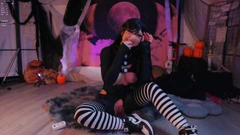 Video of a young woman in a black costume with a black and white striped mask, sitting cross-legged on a gray fur rug in a Halloween-themed room with orange pumpkins and a moon backdrop.