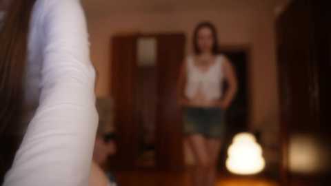 Blurry video of a woman in a white top and shorts standing in a dimly lit room with a lamp and wooden furniture.