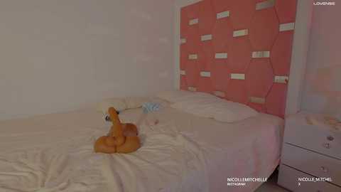 A video of a minimalist bedroom with a pink, hexagonal headboard and beige walls. A stuffed fox toy sits on a bed with white sheets.