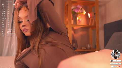 A video of a young woman with long brown hair, wearing a sheer brown top, adjusting her bra, in a warmly lit bedroom.