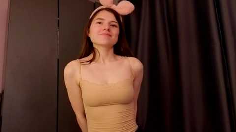 Video of a fair-skinned young woman with straight brown hair, wearing a beige camisole and bunny ears, posing against a black curtain backdrop.