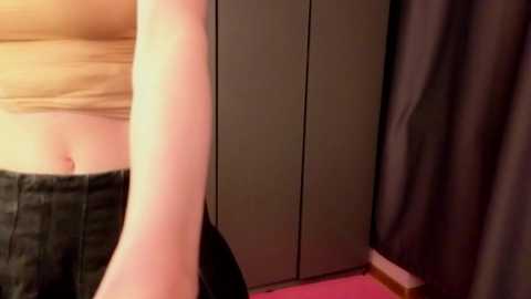 A video of a woman's lower torso and legs in a dimly lit room. She wears a beige crop top and black shorts, standing next to a dark wardrobe with red lighting casting shadows.