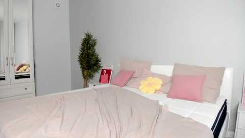 Video of a minimalist bedroom with a white bed, beige bedding, pink pillows, a yellow plush toy, and a green potted plant on a white nightstand.