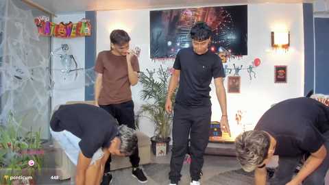 Video of three young men in casual attire, bent over, performing a push-up exercise in a modern, brightly lit room with a TV playing a video game, potted plants, and colorful decorations.