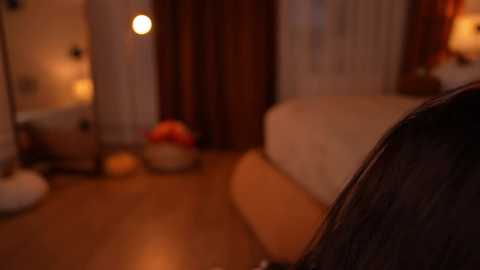 Video of a dimly-lit, cozy bedroom with a bed, beige pillows, and a warm, wooden floor. A red bowl of fruit sits on a table near a window with sheer curtains. A lamp provides soft lighting.