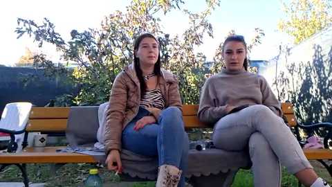 Video of two young women sitting on a wooden bench in a park. They wear casual attire, one in a striped top and jeans, the other in a beige sweater and light pants. Background features greenery and a blue sky.