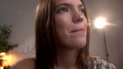 Video of a young woman with light skin, brown hair, and blue eyes, smiling slightly, wearing a gray sweater, in a dimly lit indoor setting with a spotlight in the background.