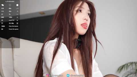 Video of an East Asian woman with long, straight brown hair, wearing a white blouse, standing in a modern, minimalist room.