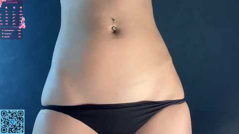Video of a light-skinned person wearing black bikini bottoms, showing navel piercing. Background features a dark wall with a QR code and calendar.
