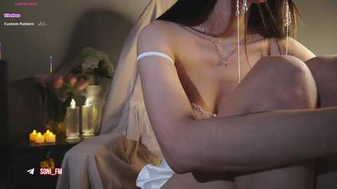 A video of a fair-skinned woman with long hair, wearing a white dress, embracing a man with a fair complexion, in a dimly lit room with candles and flowers.