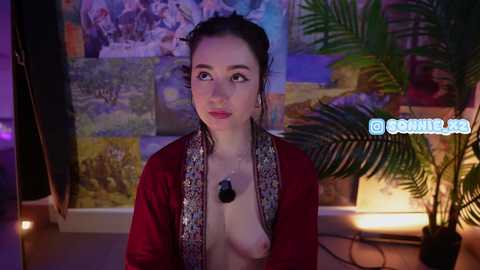 A video of a young woman with fair skin and dark hair, topless with a black necklace, standing in front of a colorful, artistic background.