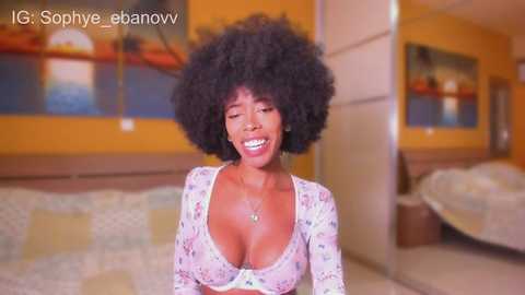 Video of a joyful, dark-skinned woman with a voluminous afro and a floral-patterned, low-cut top, sitting on a bed in a warmly lit bedroom.
