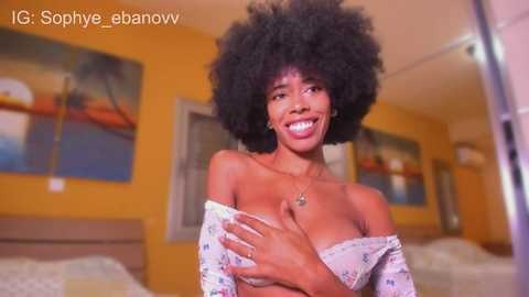 Video of a joyful, dark-skinned woman with voluminous afro, wearing a floral off-shoulder top, in a brightly lit, modern bedroom with abstract artwork on the walls.