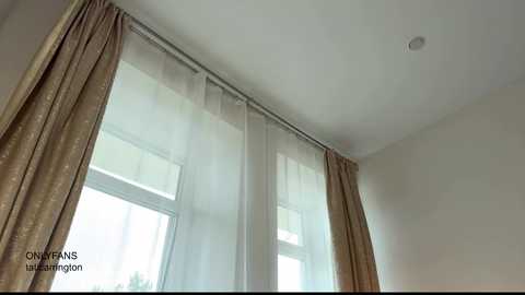 Video of a minimalist room with large, sheer white curtains and brown patterned drapes covering a window, letting soft light filter through. The ceiling has a small, circular light fixture.