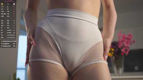 Video of a woman's lower body in white, sheer panties, hands on hips, in a modern, well-lit room with pink flowers in the background.