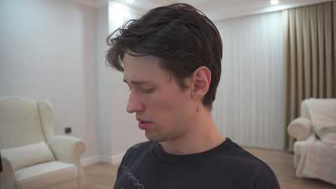 Video of a young Caucasian man with short dark hair, wearing a black t-shirt, standing in a modern, minimalist living room with beige furniture, white walls, and vertical blinds.
