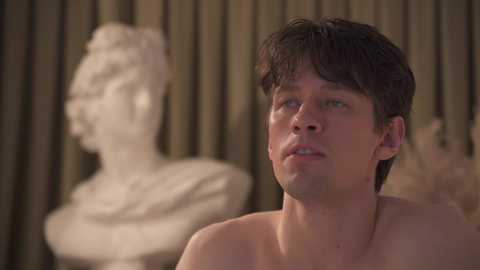 Video of a shirtless young man with short, curly brown hair, looking contemplative, standing in front of a blurred white bust statue with vertical wooden panels in the background.