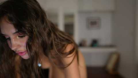 Video of a young woman with long, wavy dark hair, wearing a strapless top, leaning forward with a contemplative expression. Background features a blurred kitchen with white cabinets and a framed photo.