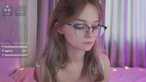 A video of a fair-skinned young woman with straight, shoulder-length brown hair, wearing glasses and a white tank top, seated indoors with soft, purple and pink lighting.