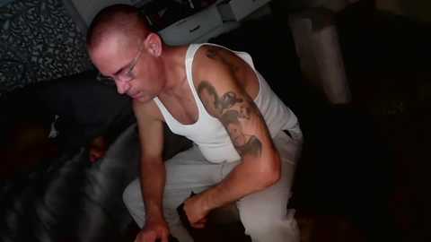 Video of a bald, muscular man with a tattooed right arm, wearing a white tank top and jeans, kneeling on a dark carpet, in a dimly-lit room with a patterned wall.