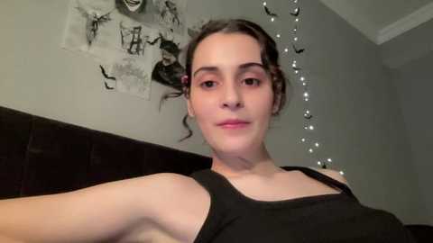 A video of a young woman with light skin, brown hair, and small breasts, wearing a black tank top, sitting on a dark couch. The background features a wall covered with black-and-white art and fairy lights.
